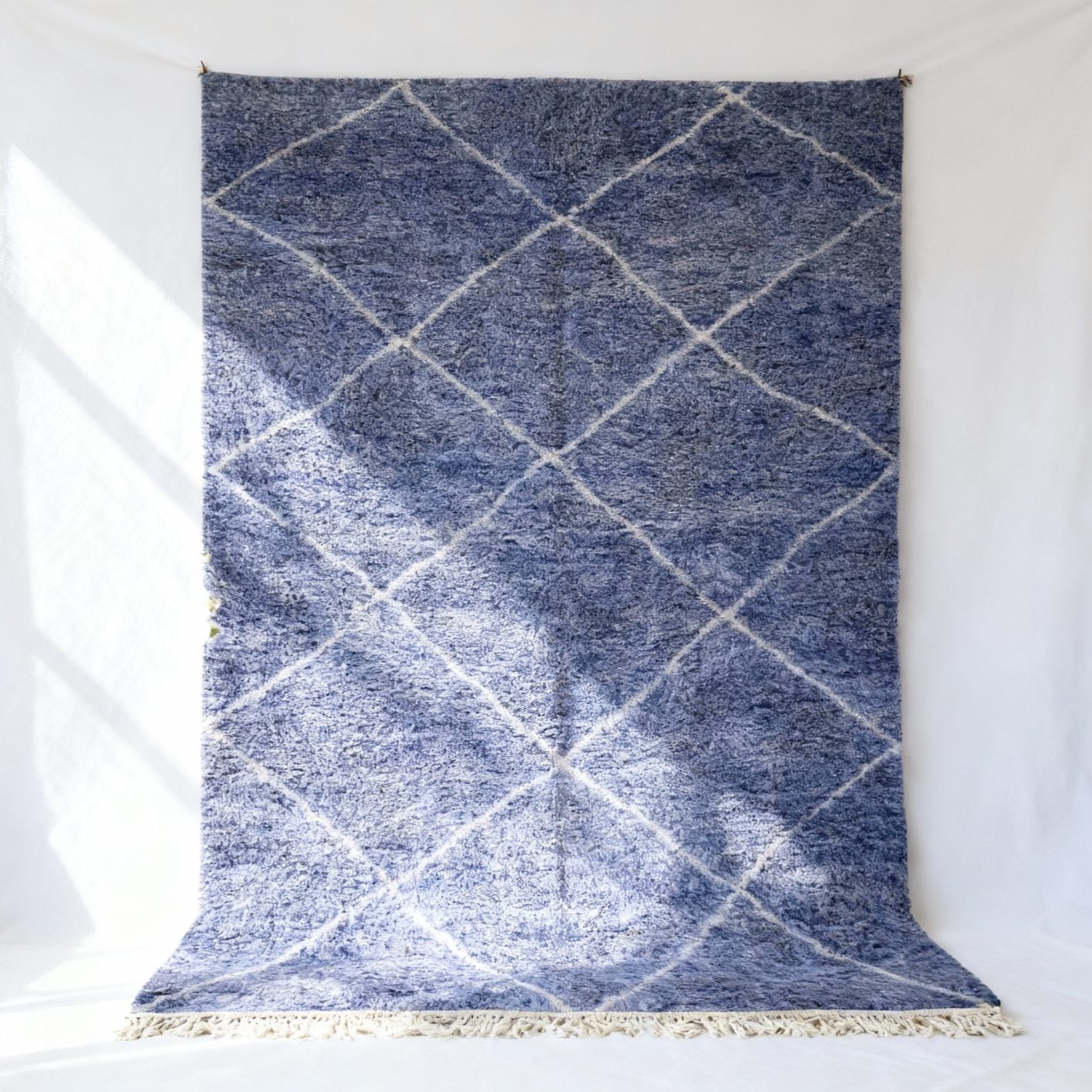 Sky blue farmhouse wool rug with white diamond lattice pattern and cotton fringe hanging on white wall Sky blue farmhouse wool rug with white diamond lattice pattern and cotton fringe hanging on white wall