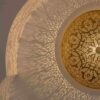Brass wall sconce projecting dramatic sunburst shadow pattern on ceiling showing detailed light rays from perforated arabesque design