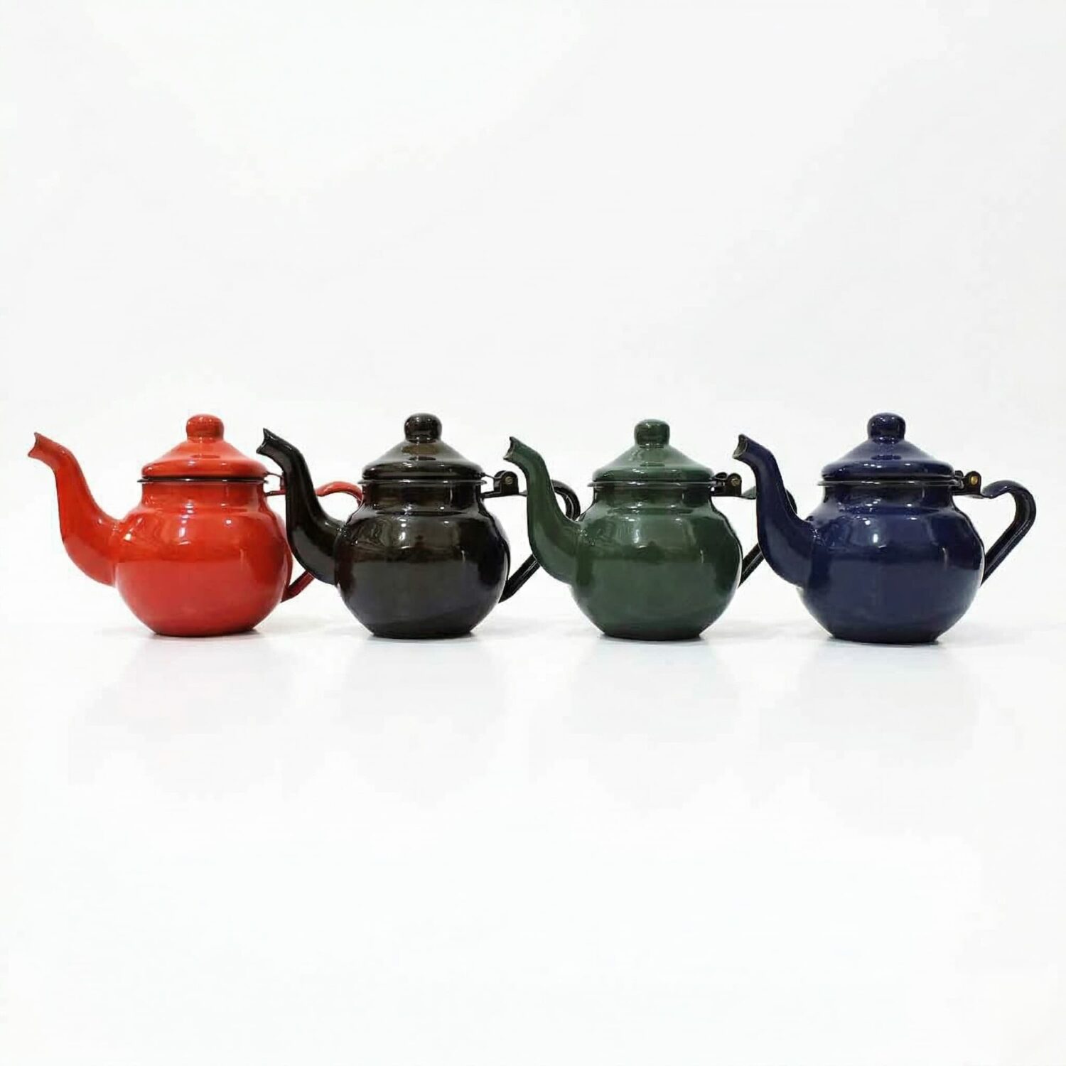 Four solid color enamel teapots in red, charcoal, green, and blue lined up on a white background showing all available c Four solid color enamel teapots in red, charcoal, green, and blue lined up on a white background showing all available colours