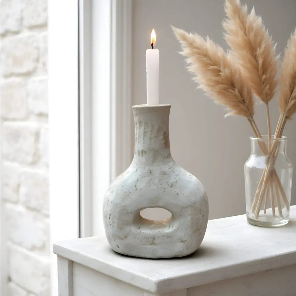 Gray ceramic candle holder with lit taper candle, handcrafted bottle design with cutout, next to pampas grass