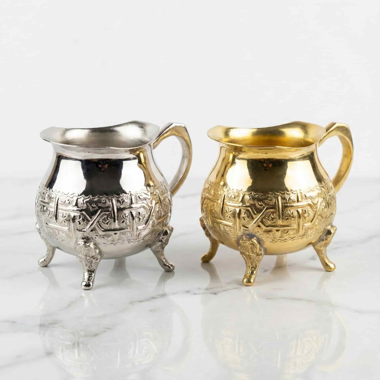 Hand-engraved mini creamer pitchers in silver and brass gold finish displayed on marble surface Hand-engraved mini creamer pitchers in silver and brass gold finish displayed on marble surface