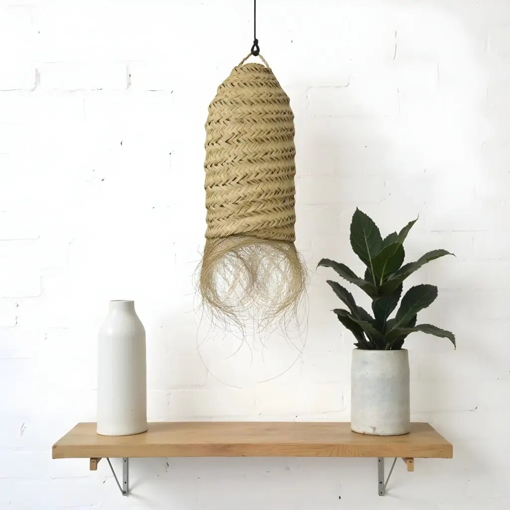Handwoven straw lighting pendant with cylindrical herringbone weave and natural fringe detail on wooden shelf