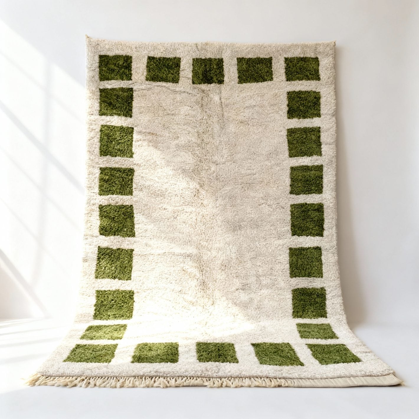 Handwoven Mrirt wool rug with emerald green geometric border on cream field, draped against white wall showing plush texture