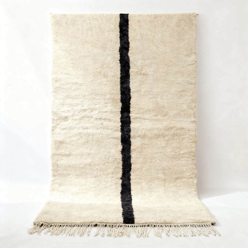 Handwoven cream wool Beni Ourain rug with minimalist single black line design, displayed on white wall Handwoven cream wool Beni Ourain rug with minimalist single black line design, displayed on white wall
