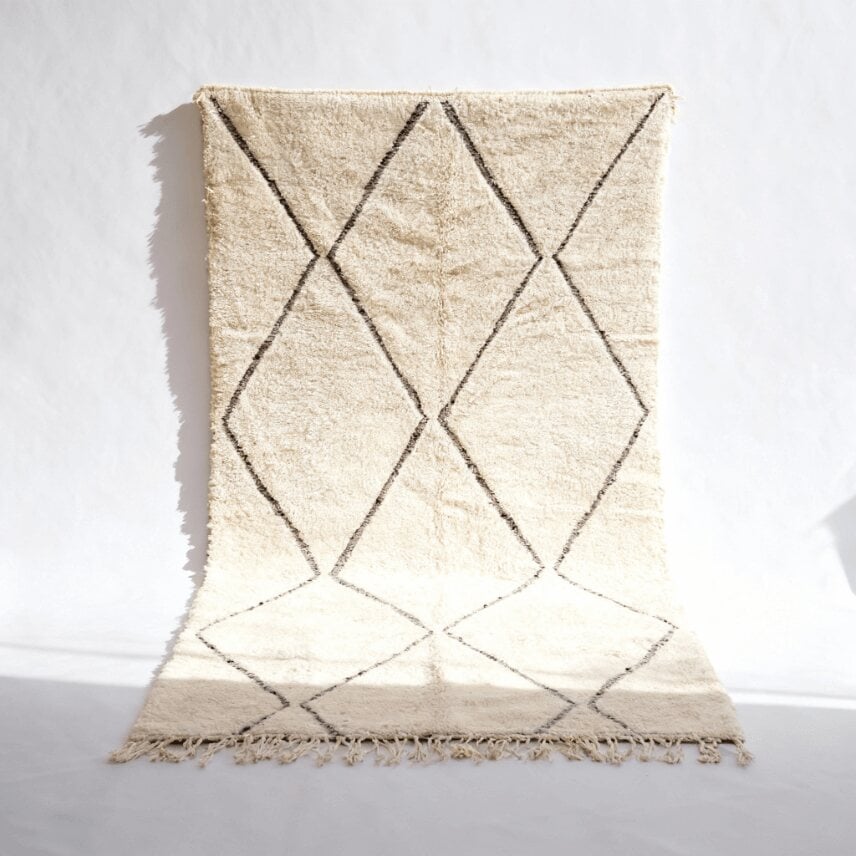 Cream and charcoal Beni Ourain wool rug with diamond pattern hanging against white wall with fringe edges Cream and charcoal Beni Ourain wool rug with diamond pattern hanging against white wall with fringe edges