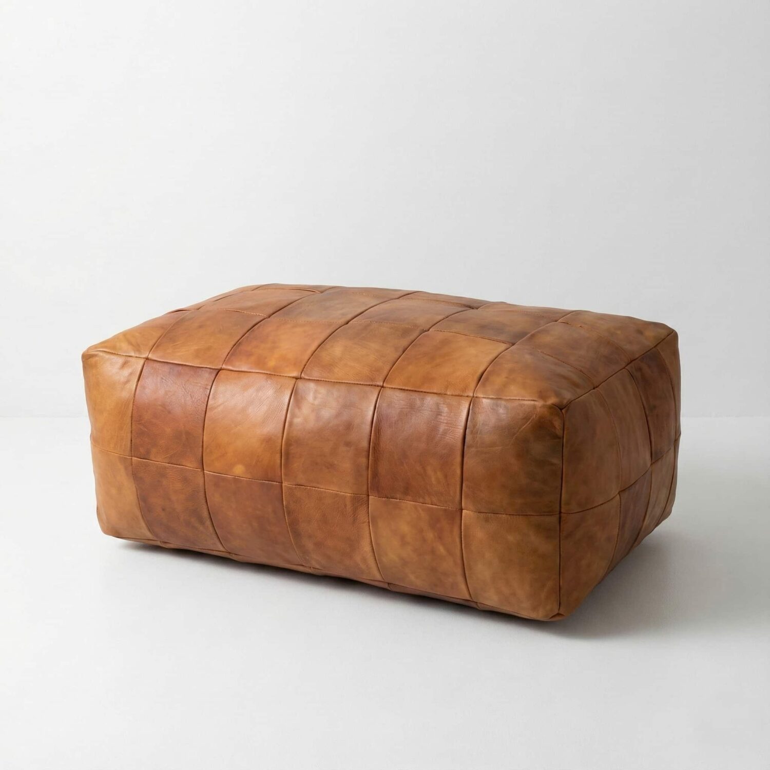 Large brown rectangular leather ottoman shown from the side with hand-stitched panel seams on a white background Large brown rectangular leather ottoman shown from the side with hand-stitched panel seams on a white background