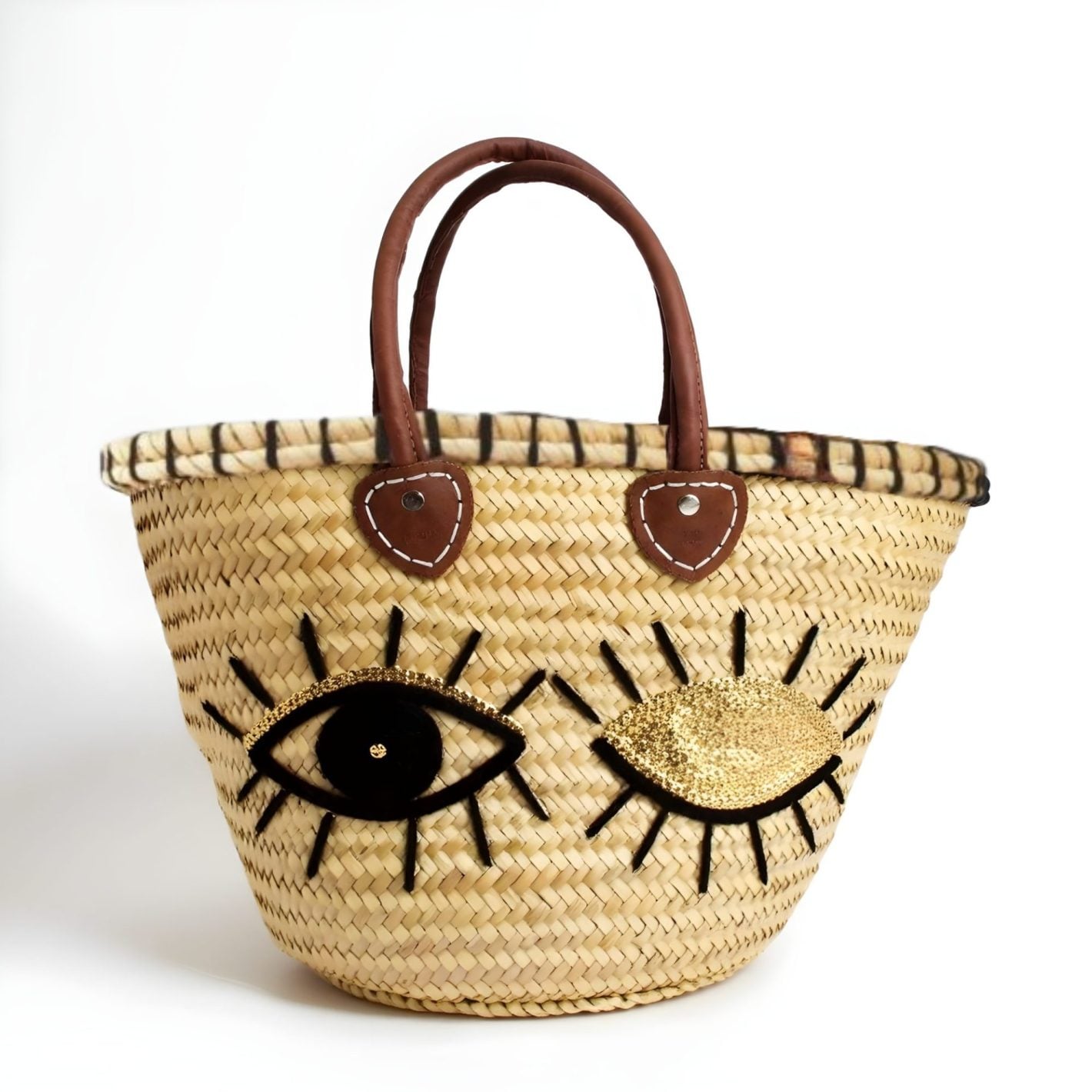Sequin straw bag with winking eye appliqué, gold sequins, brown leather handles and black whipstitch rim on white background