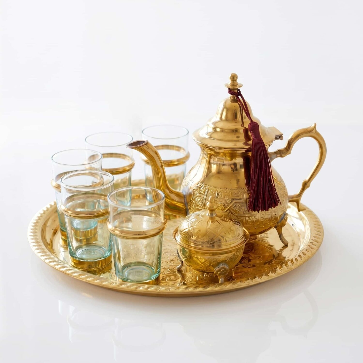 Handcrafted brass tea set 9-piece with sugar bowl, featuring an engraved teapot with a burgundy tassel, six glasses with Handcrafted brass tea set 9-piece with sugar bowl, featuring an engraved teapot with a burgundy tassel, six glasses with brass holders, and a matching serving tray isolated on a cl