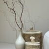 Two Moroccan lidded seagrass baskets with white trim and wool pom pom lids beside a ceramic vase