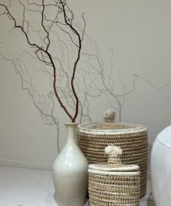 Two Moroccan lidded seagrass baskets with white trim and wool pom pom lids beside a ceramic vase