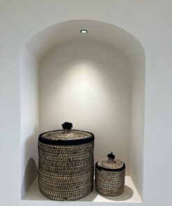 Two Moroccan lidded seagrass baskets with black stitching and black pom pom lids in a lit wall niche
